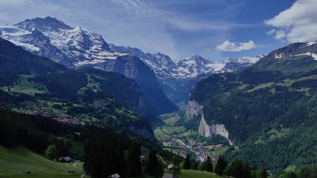 Sweeping drone panorama of Lauterbrunnen Valley, Swiss Alps, with alpine villages, steep cliffs, green slopes and snowcapped peaks under a blue sky.