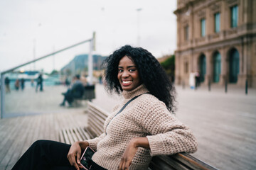 Fototapeta premium Smartphone application chat network user online shopping browsing. Confident smiling African American woman sitting on city bench looking at camera holding mobile device, inclusion real people