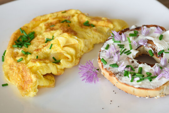 Close up of a white plate with an omelette and bagel topped with cream cheese, garnished with fresh green chives and purple chive blossom flowers