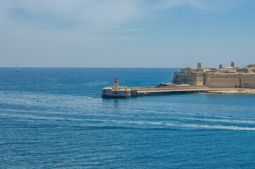 Fototapeta premium Red lighthouse on stone breakwater at harbor entrance with ancient fort sailboats and blue Mediterranean sea on sunny summer day