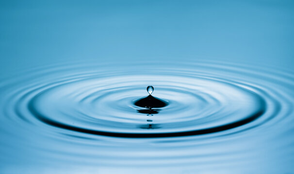Close up of a blue water drop macro creating concentric ripples on a calm water surface. Abstract liquid background symbolizing purity, freshness, balance and clean water