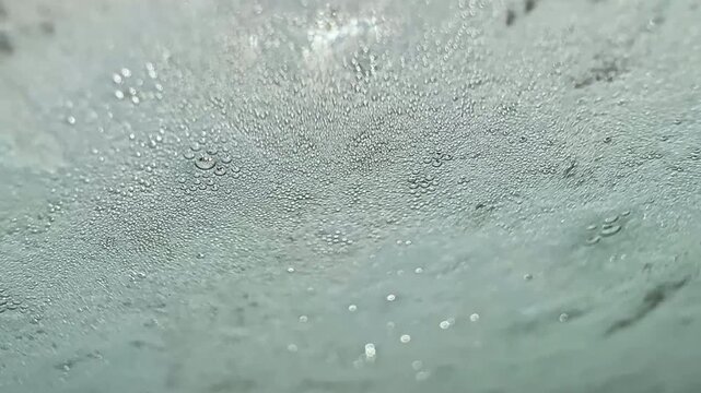 Water droplets gather on a transparent surface. The droplets vary in size and create a design as rain falls outside. Light reflects in the water, adding to the view.