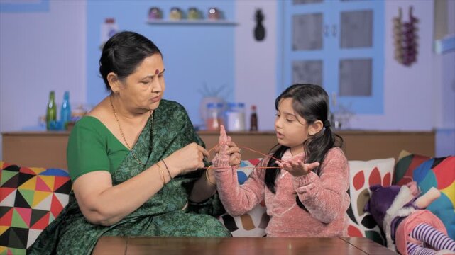 Grandmother teaching a little child to make a pattern on fingers using thread. Elderly lady playing cat's cradle game with her granddaughter while sitting indoors - relationship and bonding