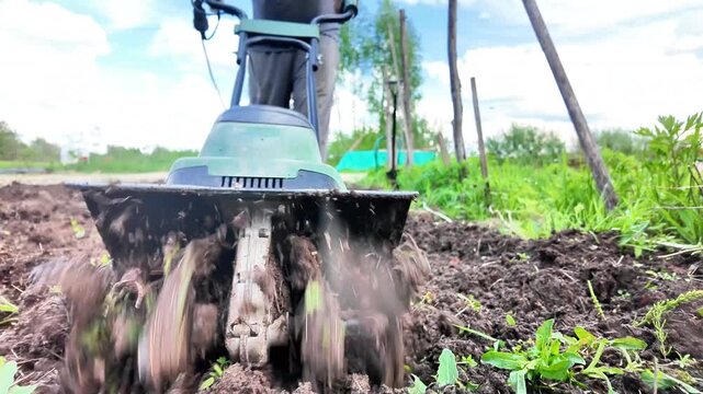 Ground-level close-up of a rotary cultivator breaking through wet garden soil enriched with organic compost. Large clumps of earth fly toward the camera as the machine works through dense ground