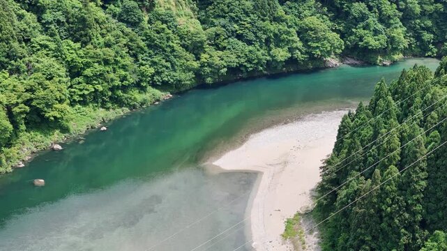 Aerial view of a clear river winding through dense green forest. Shallow sandy patches contrast with deep turquoise water. Lush trees line both sides of the river's gentle curve