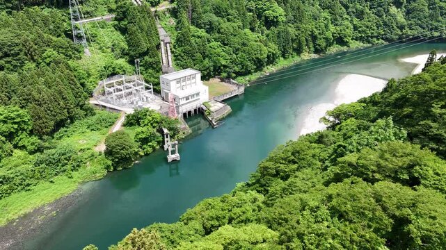 Aerial view of river flowing through green forested hills. Concrete dam structure stands beside turquoise water. White water rushes near the dam's intake