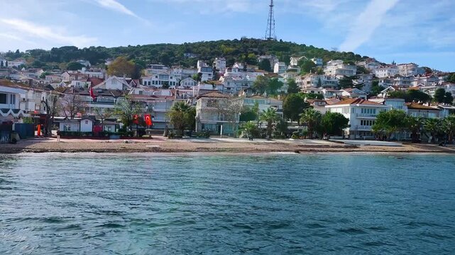 Beautiful scenic view of Kinaliada Island coastline featuring a sandy beach, waterfront houses, and green hills under a bright blue sky, captured from the sea in Princes Islands, Istanbul, Turkey