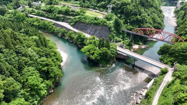River winds through lush green hills. Concrete bridge spans rushing water below. Red metal arch bridges stand tall, sturdy. Riverside paths wind near rocks and trees