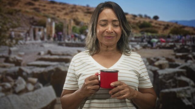 Woman holding a red cup with eyes closed in front of an ancient building ruins, hands wrapped around enamel mug; calm reflection.