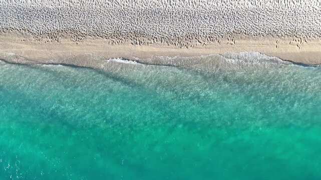 Aerial view of clear turquoise sea water gently lapping against a sandy beach, showcasing intricate patterns of footprints in the sand along the shoreline. Ocean waves at coastline. Summer vacation