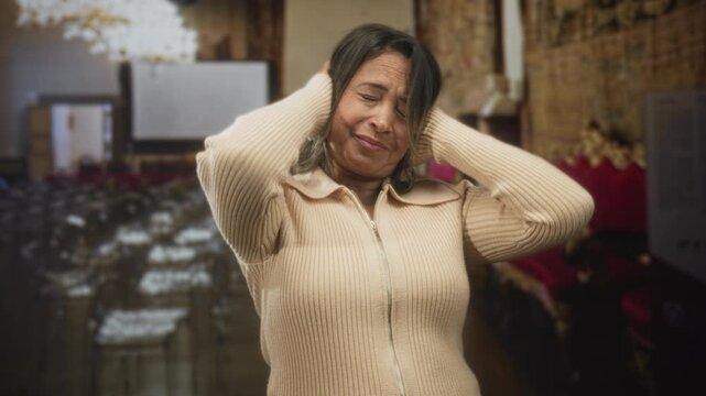 Woman presses hands to ears and winces, standing among rows of chairs and gilded seating in an ornate hall building; distress sensory overload.