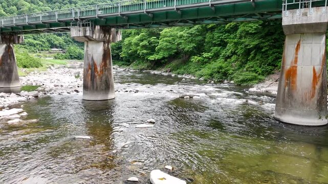 Green bridge spans river, supported by stone pillars. Rust stains mark pillars where water meets concrete. Riverside stones glow under gentle current's flow