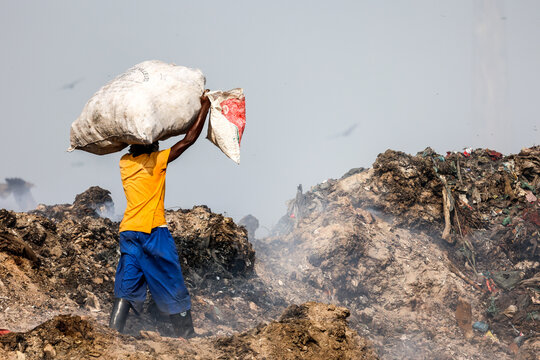 Aminbazar, Bangladesh - 21 February 2026: View of a worker carrying a heavy sack on his head through the Aminbazar landfill amidst piles of waste and rising smoke.
