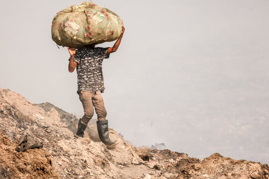 Aminbazar, Bangladesh - 21 February 2026: View of a worker carrying a large sack of waste on his head while walking across the Aminbazar landfill site in a hazy environment.