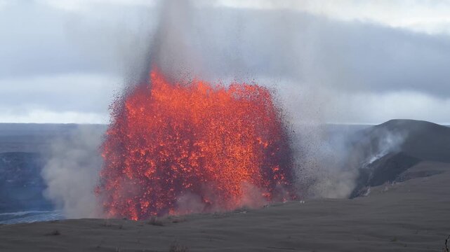 View of a massive lava fountain erupting from Halemaumau Crater with thick smoke and steam rising into a cloudy sky in Hawaii County, Hawaii, United States.