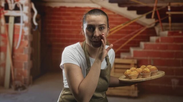 Woman holding muffins on a wooden tray in a bakery, smiling and calling out while gesturing near her mouth, with brick wall, staircase and wooden crates behind her; cozy hospitality.