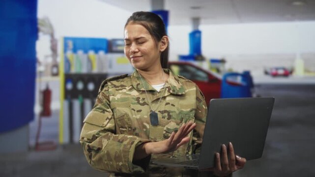Woman soldier holding laptop with palm raised, glancing at screen, wearing dogtags and camouflage uniform in a building forecourt; duty caution resilience.