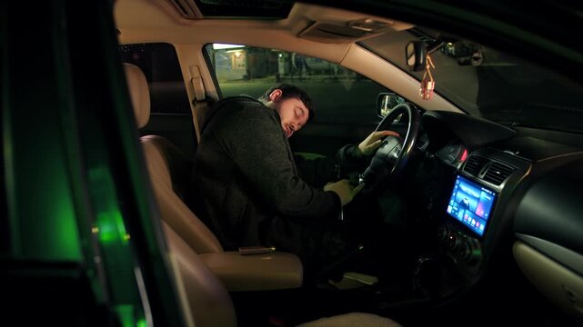 Unaware sleepy drunk caucasian driver is in stationary parked automobile with open door on empty lot at night. An intoxicated man breaks the law and fell asleep in a bright salon of vehicle in city