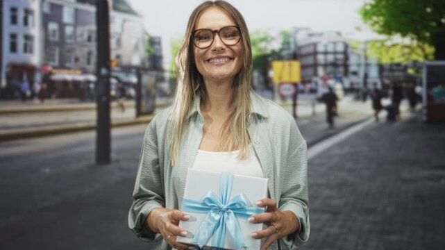 Woman holding gift box with hands on a busy street smiling and presenting a blue ribbon wrapped package; warm generosity joy.