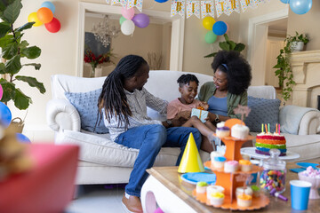 Obraz premium African American family sitting on white sofa celebrating child opening wrapped gift in living room