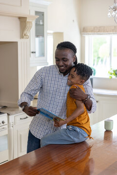 African American father and child reading blue book while sitting on wooden counter in kitchen