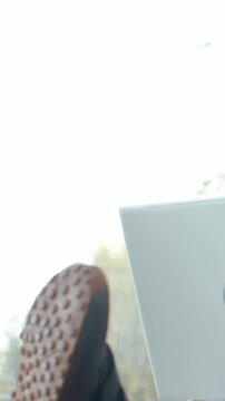 Vertical video: Propping person's foot near laptop by window, showing dark sandal sole, copy space