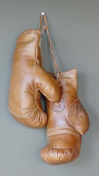 Vertical video: Hanging brown leather boxing gloves swinging on gray wall, showing laces, hook