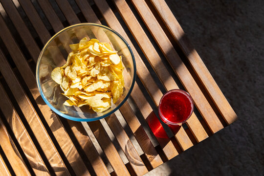 Clear glass bowl potato chips sitting on patio slat table beside short red tumbler casting stripes