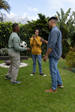 Diverse teenage males standing on backyard lawn holding soccer ball, clapping beside stone planter