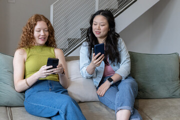 Diverse female friends sitting on sofa in living room comparing smartphones with visible smartwatch © wavebreak3