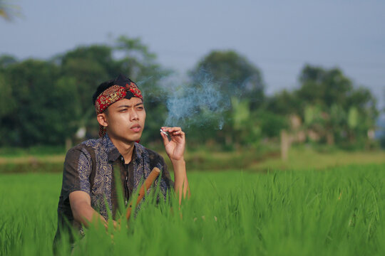 An Indonesian Asian man wearing a traditional batik shirt and udeng headpiece, relaxing while holding a cigarette in a lush green rice field under a clear blue sky.