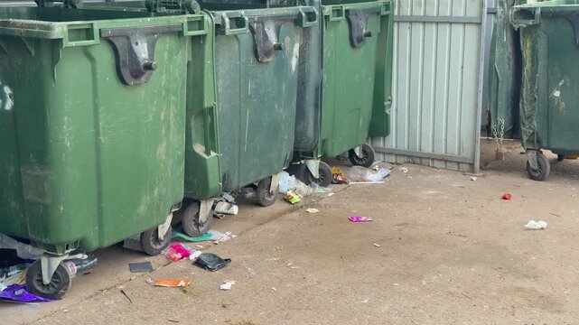Green dumpsters with scattered trash under bright summer sunlight, gravel foreground, worn metal bins, colorful packaging