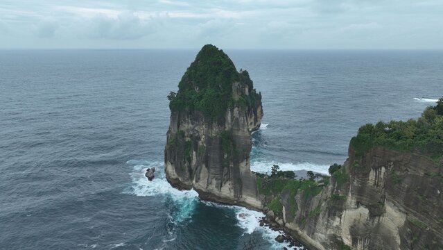 Aerial view of the iconic sea stack at Pantai Pangasan with waves crashing against the steep cliffs under an overcast sky in Kecamatan Kebonagung, Jawa Timur, Indonesia.