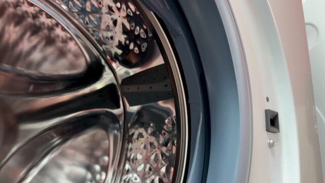 Close-up steel drum washing machine door showing gleaming stainless interior with perforated pattern, open rubber gasket, visible