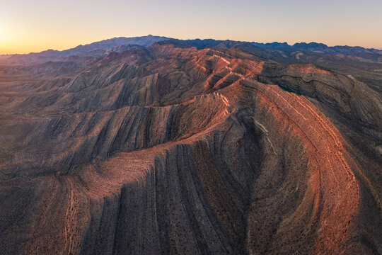Aerial view of the geological folds and rugged ridges of Potosi Mountain glowing under the warm light of sunset in Clark County, Nevada, United States.