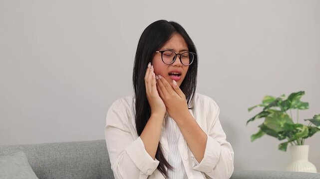 Young Asian woman sitting on sofa while touching her sore jaw and suffering from stubbing toothache