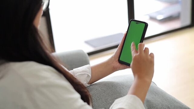 Back view of Young Asian Woman sit use digital tech cell phone open green screen mock up at home