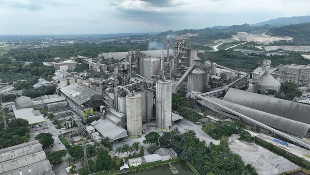 Aerial view of a large industrial cement plant with tall silos, complex machinery, and conveyor belts surrounded by lush vegetation and hills Klapanunggal, West Java, Indonesia.