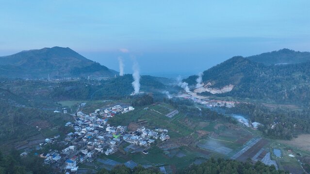 Aerial view of the Dieng Plateau featuring the village of Kejajar surrounded by volcanic mountains and geothermal steam plumes under a twilight sky in Kejajar, Central Java, Indonesia.