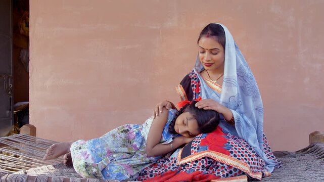 Medium shot - Indian village woman making her little daughter sleep on her lap. Medium shot of a mother-daughter duo sitting on a woven cot during a hot summer day in a village - parenting