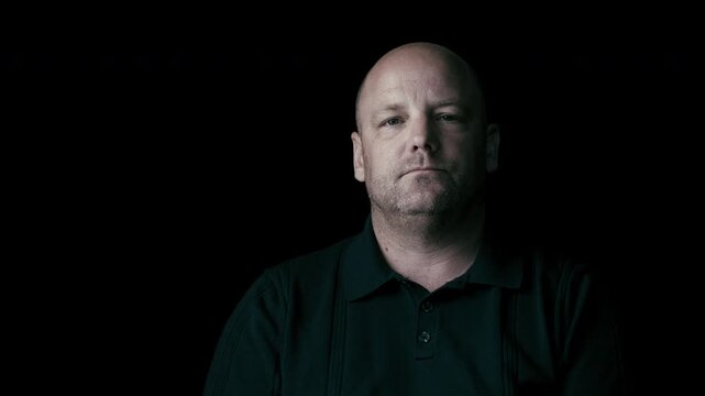 White middle-aged man in front of a plain background. Dressed in a black collared shirt with shadow casting on the person's right side of the face. 