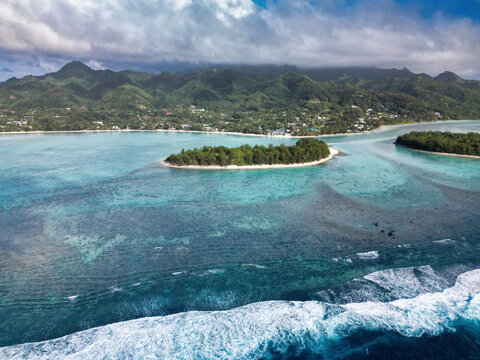 Aerial view of the turquoise waters of Muri Lagoon with a lush green islet and coral reefs against the backdrop of volcanic mountains Cook Islands.