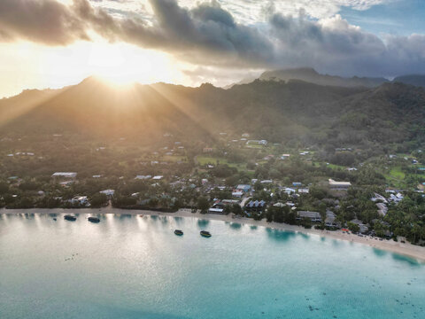 Aerial view of a tropical coastline with turquoise water, sandy beaches, and lush green mountains under a dramatic sunset with sun rays in Cook Islands.
