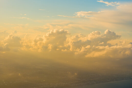 Massive cumulus cloud formations illuminated by warm golden sunlight floating above a hazy distant coastal city landscape.