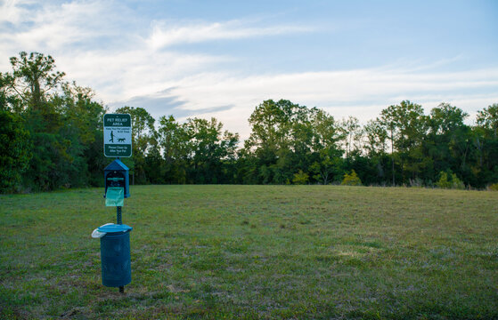 Green metal pet relief area sign and a blue plastic dog waste bag dispenser standing securely on a large open grassy park field.