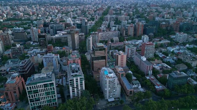 Retreating reveal over Providencia rooftops exposing tree-lined boulevard cutting through vast urban grid fading to city limits at blue hour. Santiago, Chile