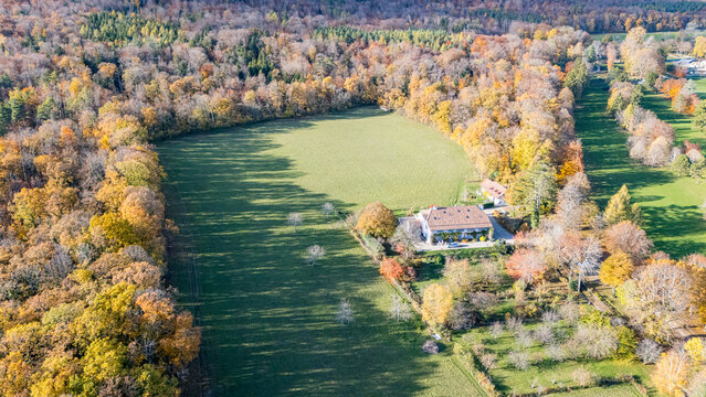 Aerial view of a traditional farmhouse at the edge of a vast green meadow surrounded by a dense forest with autumn foliage in the Jura mountains La Rippe, Vaud, Switzerland.