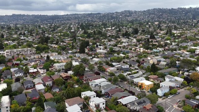 Berkeley residential neighborhood with tree-lined streets and single-family homes with solar panels, dense wooded hills rising behind under cloudy sky in spring. California, USA. Aerial flyover.