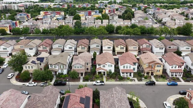 Two-story suburban homes with terracotta rooftops flanking wide boulevard, colorful townhouses with solar panels and parked cars on both sides under sunny sky. California, USA. Aerial top down.