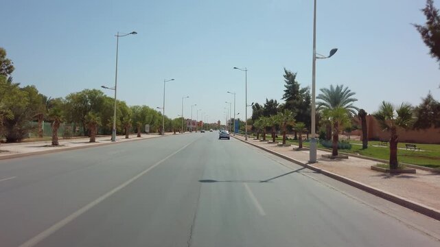 In Errachidia's Ksar es-Souk, cars move along a city road lined with buildings, storefronts, palm trees and street lamps, capturing a typical urban scene in this Moroccan desert community.